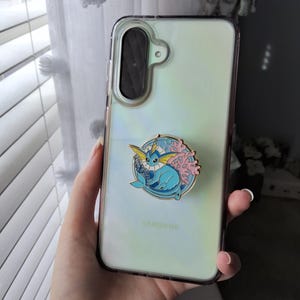 Puede incluir: Una funda transparente para tel&eacute;fono Samsung con panel trasero iridiscente y un agarre circular de esmalte. El agarre muestra un personaje Vaporeon azul rodeado de coral rosa y olas de agua azules, con un borde de metal en tono dorado.
