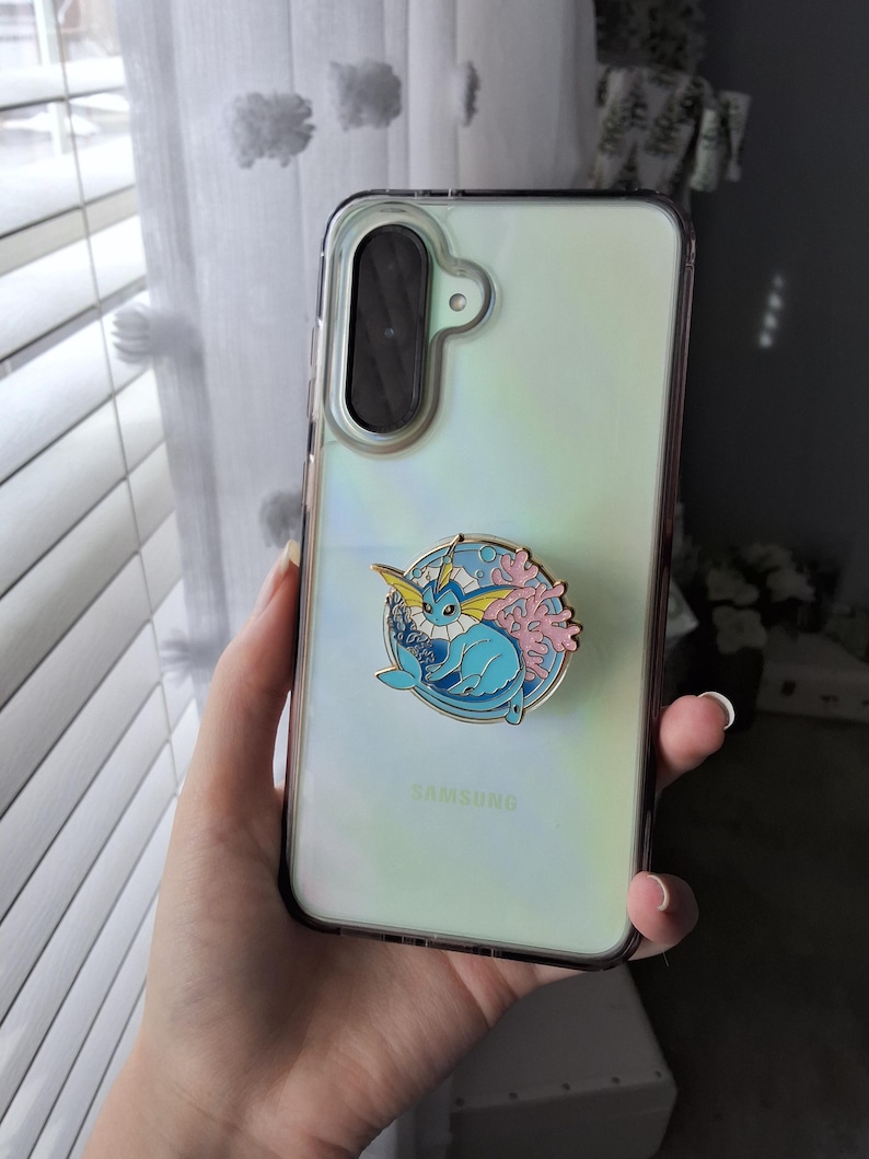 Puede incluir: Una funda transparente para tel&eacute;fono Samsung con panel trasero iridiscente y un agarre circular de esmalte. El agarre muestra un personaje Vaporeon azul rodeado de coral rosa y olas de agua azules, con un borde de metal en tono dorado.