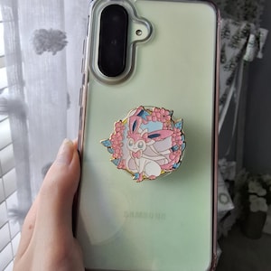 Puede incluir: Una funda transparente para smartphone con fondo verde p&aacute;lido y un soporte circular de esmalte para tel&eacute;fono en el centro. El soporte muestra un personaje de criatura rosa y blanco rodeado por una corona de flores de cerezo rosas y peque&ntilde;as hojas azules.