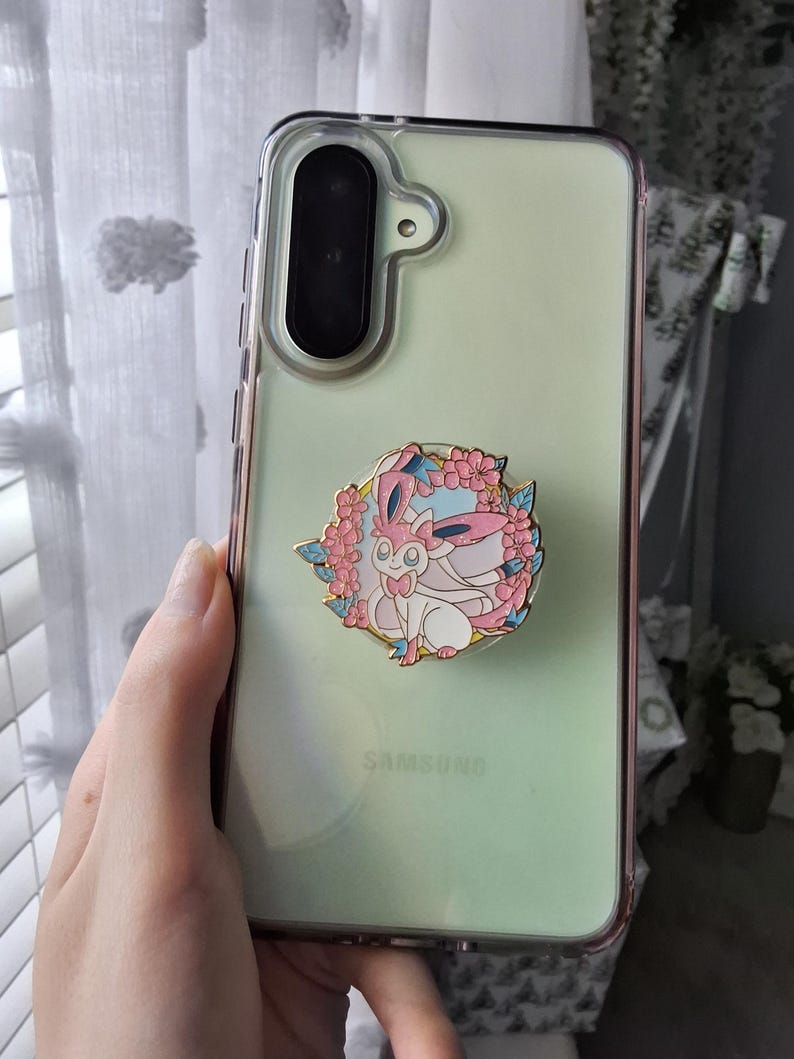 Puede incluir: Una funda transparente para smartphone con fondo verde p&aacute;lido y un soporte circular de esmalte para tel&eacute;fono en el centro. El soporte muestra un personaje de criatura rosa y blanco rodeado por una corona de flores de cerezo rosas y peque&ntilde;as hojas azules.