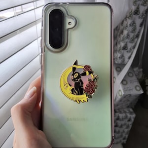 Puede incluir: Una funda transparente para smartphone con fondo verde claro y un soporte circular que muestra una criatura negra con anillos amarillos, una luna creciente amarilla con cara y rosas rojas. La palabra SAMSUNG est&aacute; impresa en la funda.