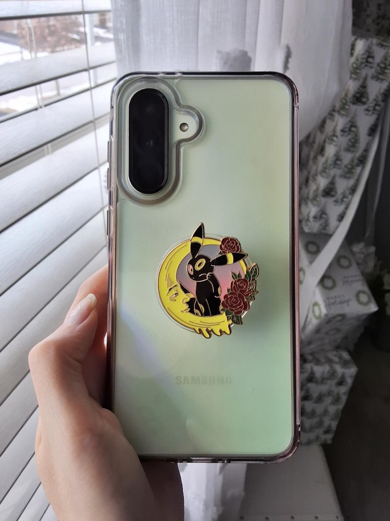 Puede incluir: Una funda transparente para smartphone con fondo verde claro y un soporte circular que muestra una criatura negra con anillos amarillos, una luna creciente amarilla con cara y rosas rojas. La palabra SAMSUNG est&aacute; impresa en la funda.