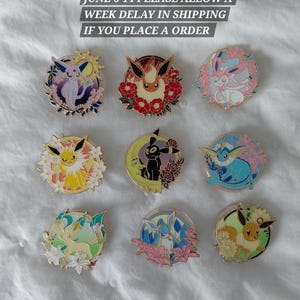 Op de afbeelding: Een collectie van negen ronde emaille pins met diverse gestileerde wezens omringd door florale en hemelse motieven in levendige kleuren. De pins tonen personages waaronder Espeon, Flareon, Sylveon, Jolteon, Umbreon, Vaporeon, Leafeon, Glaceon en Eevee.