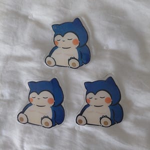 Peut inclure: Trois autocollants transparents représentant un personnage de dessin animé Snorlax en position assise. Le Snorlax a une fourrure bleue, un ventre crème et des joues oranges. Les autocollants sont sur une surface blanche texturée.