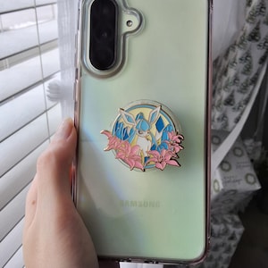 Puede incluir: Una funda transparente para tel&eacute;fono Samsung con un reverso iridiscente p&aacute;lido y un agarre circular de esmalte en el centro. El agarre presenta una ilustraci&oacute;n de una criatura azul claro y blanca rodeada de flores de lirio rosa y cristales azules.