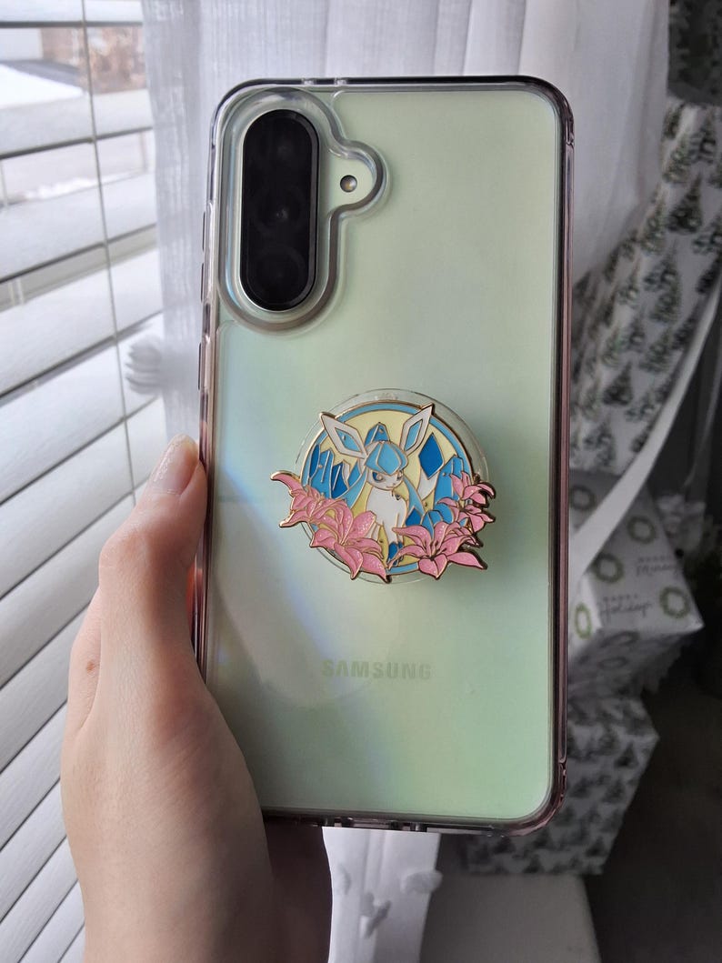 Puede incluir: Una funda transparente para tel&eacute;fono Samsung con un reverso iridiscente p&aacute;lido y un agarre circular de esmalte en el centro. El agarre presenta una ilustraci&oacute;n de una criatura azul claro y blanca rodeada de flores de lirio rosa y cristales azules.