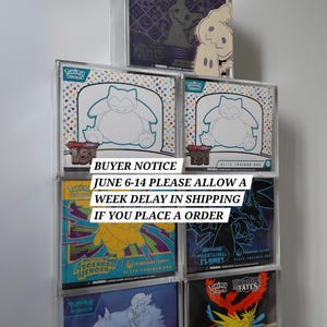 May include: A collection of six Pokémon Elite Trainer Boxes displayed in clear acrylic protective cases. The boxes feature various designs including Snorlax, Mimikyu, and other characters from the Scarlet and Violet series, stacked in a pyramid arrangement.