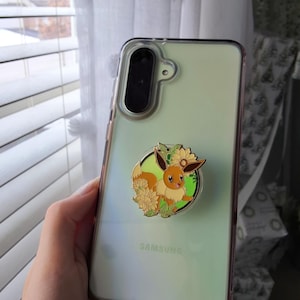 Puede incluir: Una funda transparente para tel&eacute;fono Samsung con un soporte circular que presenta una ilustraci&oacute;n esmaltada de un personaje Eevee marr&oacute;n rodeado de girasoles amarillos y hojas verdes. La funda tiene un tono verde claro y un m&oacute;dulo de c&aacute;mara negro.