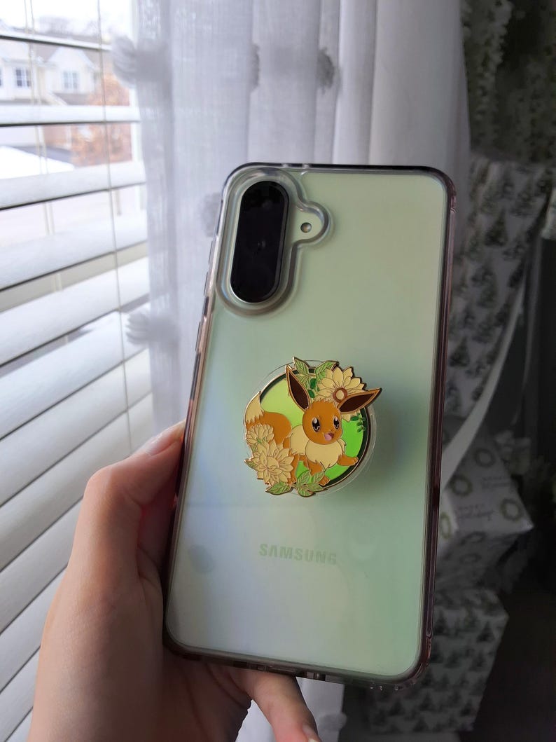 Puede incluir: Una funda transparente para tel&eacute;fono Samsung con un soporte circular que presenta una ilustraci&oacute;n esmaltada de un personaje Eevee marr&oacute;n rodeado de girasoles amarillos y hojas verdes. La funda tiene un tono verde claro y un m&oacute;dulo de c&aacute;mara negro.