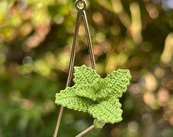 May include: A gold triangle pendant necklace with a green crocheted flower charm. The necklace is hanging from a black cord.