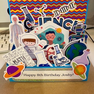 May include: A handmade birthday card with a science theme. The card features a blue box with various science-related stickers, including a microscope, astronaut, and planet. The text "SCIENCE" is prominently displayed, and the message "Happy 8th Birthday Joshy!" is written on the front.
