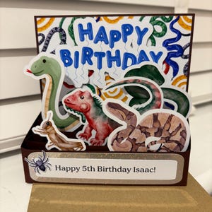 May include: A pop-up birthday card with illustrations of a green snake, a red dinosaur, and a brown snake. The card displays the words "Happy Birthday" in blue balloon letters and "Happy 5th Birthday Isaac!" on a banner.