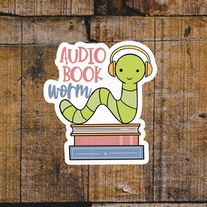 May include: A sticker featuring a cartoon worm wearing headphones, atop a stack of books. The sticker reads "AUDIO BOOK worm" in pink and blue text. The worm is green with yellow accents.
