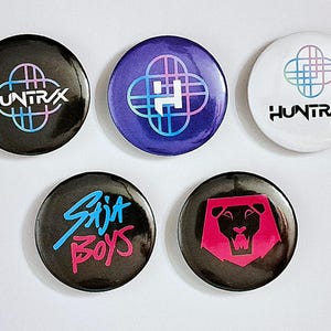 May include: Five circular badges with different designs. The badges display the text "HUNTRAX" and "Saja Boys", a lion graphic, and abstract shapes. The badges are black, purple, and white with colourful accents.