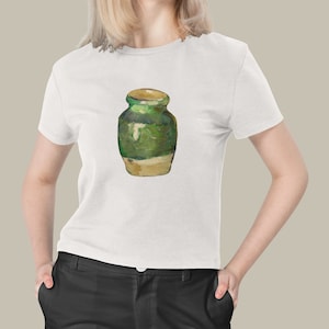 Op de afbeelding: Wit T-shirt met een aquarel schilderij van een groen en beige pot. De pot heeft een afgeronde vorm met een deksel en een lichtere basis. Het kunstwerk is gecentreerd op het shirt.