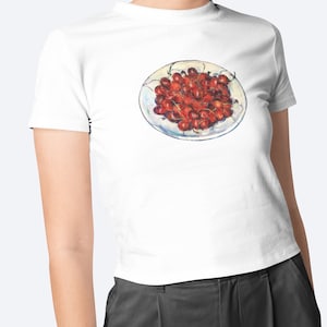 Op de afbeelding: Wit T-shirt met een schilderij van een bord met rode kersen. De kersen hebben groene steeltjes en zijn geschilderd in een realistische stijl. Het T-shirt heeft een ronde hals.