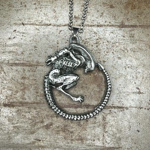 Könnte beinhalten: Eine silberfarbene Halskette mit Anhänger, die ein detailliertes Xenomorph-Design zeigt. Der Anhänger zeigt die Kreatur in einer kreisförmigen Pose, mit einer strukturierten Kette. Die Halskette ist aus glänzendem Metall.
