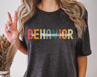 以下が含まれることがあります： カラフルなレトロな文字で「BEHAVIOR is communication」と書かれたダークグレーのヘザーTシャツです。