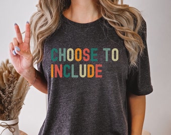 以下が含まれることがあります： レトロな虹色のカラーリングで「CHOOSE TO INCLUDE」という文字が書かれたダークグレーのヘザーTシャツです。