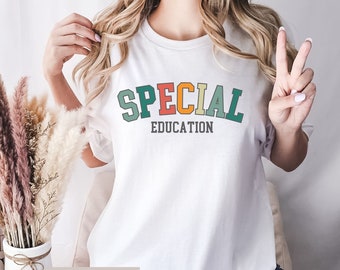 Puede incluir: Camiseta blanca con el texto "SPECIAL EDUCATION" en una fuente de estilo retro con letras de colores. La camiseta está siendo modelada por una persona que lleva pantalones vaqueros azules.