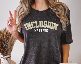 以下が含まれることがあります： 黄金色で「INCLUSION MATTERS」と書かれたダークグレーのヘザーTシャツ。テキストは大学風のフォントです。