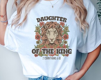 May include: White t-shirt with a graphic of a lion's head surrounded by flowers. The text "Daughter of the King" and "2 Corinthians 6:18" are printed on the shirt.