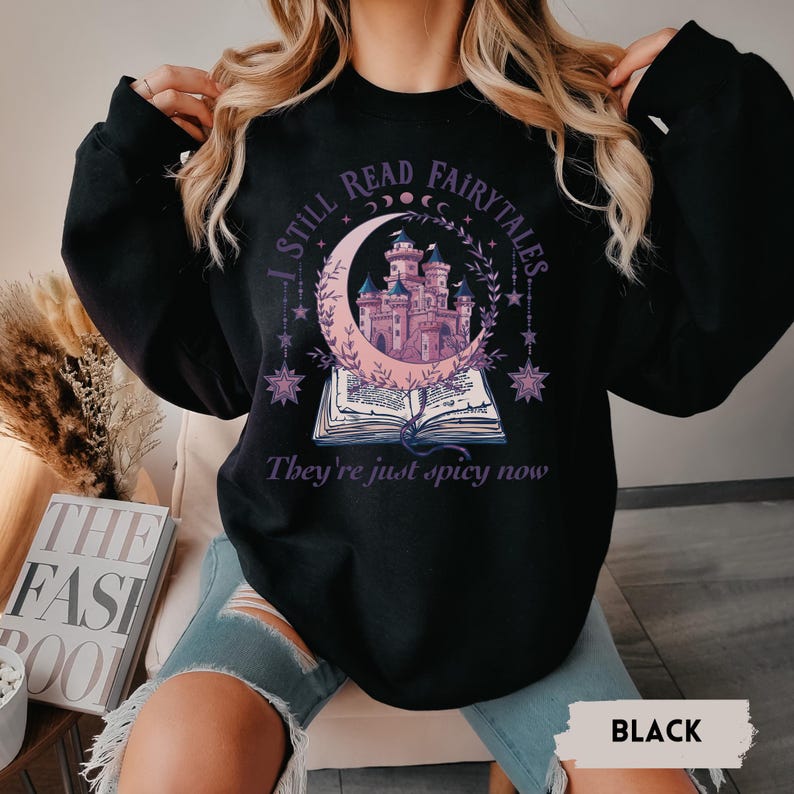 May include: Black sweatshirt with a graphic design featuring a pink castle inside a crescent moon, an open book, and the text "I STILL READ FAIRYTALES. They're just spicy now."