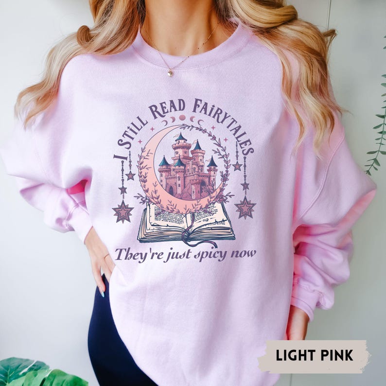 May include: Light pink sweatshirt with a graphic design featuring a castle inside a crescent moon, stars, and an open book. The text reads "I STILL READ FAIRYTALES" and "They're just spicy now."