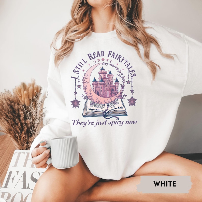 May include: White sweatshirt with a graphic featuring a pink castle inside a crescent moon, surrounded by stars and the text "I STILL READ FAIRYTALES, They're just spicy now."