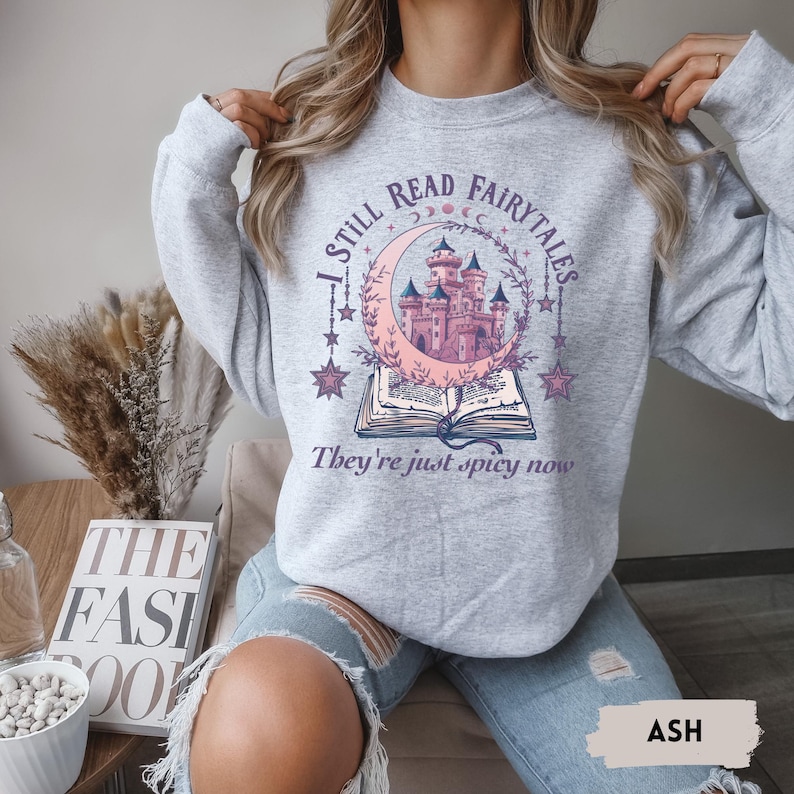 May include: Ash gray sweatshirt with a whimsical design. The graphic features a pink castle inside a crescent moon, surrounded by stars and a floral wreath, above an open book. Text reads "I STILL READ FAIRYTALES. They're just spicy now."
