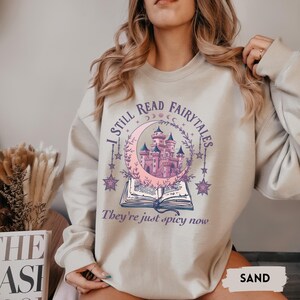 May include: Sand-colored sweatshirt with a whimsical design. The graphic features a pink castle inside a crescent moon, surrounded by stars, and an open book. Text reads "I STILL READ FAIRYTALES, They're just spicy now."