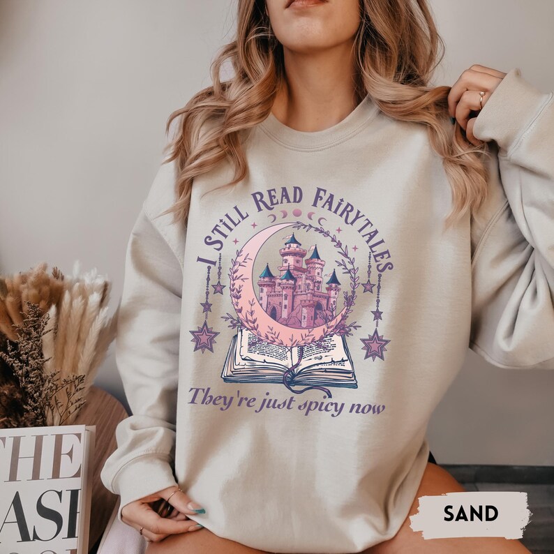May include: Sand-colored sweatshirt with a whimsical design. The graphic features a pink castle inside a crescent moon, surrounded by stars, and an open book. Text reads "I STILL READ FAIRYTALES, They're just spicy now."