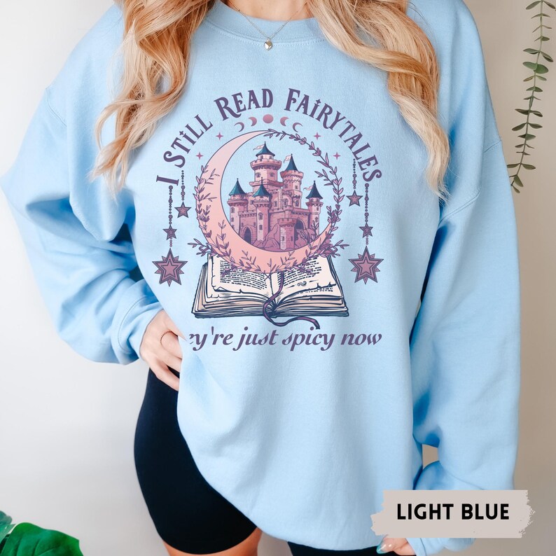 May include: Light blue sweatshirt with the text "I STILL READ FAIRYTALES" above a graphic of a castle inside a crescent moon, and an open book. The text "they're just spicy now" is below the book. The words "LIGHT BLUE" are at the bottom.