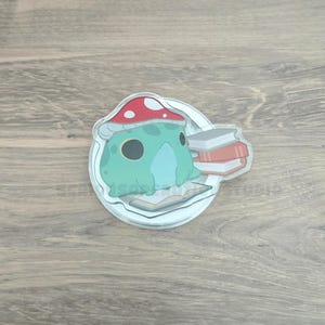 May include: A whimsical sticker featuring a teal frog wearing a red mushroom cap, sitting on a book. A stack of books is to the right. The sticker has a white border and is on a wooden surface.
