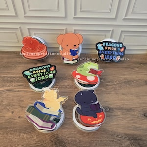 May include: A collection of acrylic drink coasters featuring various cartoon characters and designs. The coasters include a bear reading a book, a frog on a mushroom, and a black cat on a pumpkin. Some coasters have the text "DRAGONS SPICE & EVERYTHING ICED".