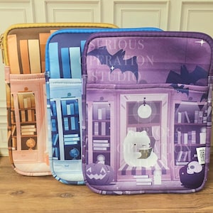 May include: Three zippered tablet cases with unique designs. One is brown with a library theme, another is blue with a similar theme, and the third is purple with a Halloween-themed illustration and the text "SERIOUS OPERATION STUDIO".