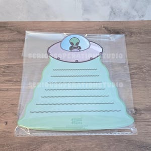 May include: A light green notepad shaped like a UFO, with a blue and gray alien design. The notepad has horizontal lines for writing and is packaged in a clear plastic sleeve. The text "SERIOUS OPERATION STUDIO" is printed on the notepad.