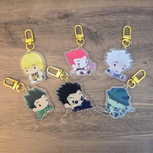 May include: A collection of seven acrylic keychains featuring anime-style character designs. Each keychain has a gold-colored clasp and depicts a different character with distinct hair colors and outfits. The keychains are arranged on a wooden surface.