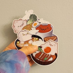 Puede incluir: Un conjunto de cuatro pegatinas con adorables gatos de dibujos animados disfrutando de varios platos japoneses. Las pegatinas están impresas en papel blanco con un acabado brillante. Los platos incluyen ramen, un tazón de sopa con tofu, un tazón de arroz con un huevo y un tazón de arroz con carne.