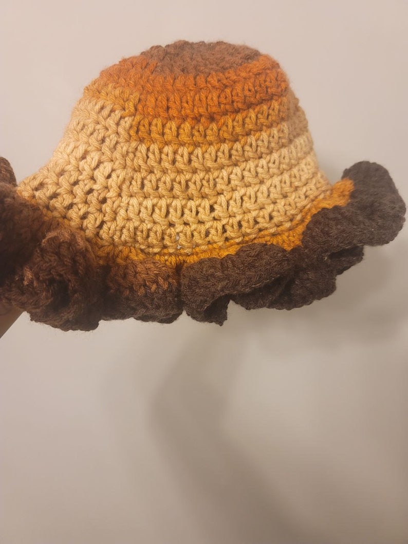 Pu&ograve; includere: Un cappello a secchiello all'uncinetto con un design a ombre di marrone, arancione e beige. Il bordo del cappello &egrave; decorato con volant in crochet marroni.