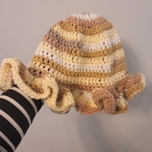 Pu&ograve; includere: Un cappello a secchiello all'uncinetto con un bordo arricciato. Il cappello &egrave; a righe in tonalit&agrave; beige, giallo e bianco.