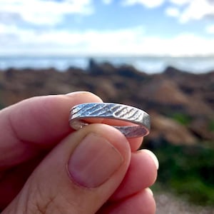 Könnte beinhalten: Ein silberner Ring mit strukturierter Oberfläche, gehalten in einer Hand. Das Design des Rings weist ein Muster aus diagonalen Linien auf. Der Hintergrund zeigt eine verschwommene Ansicht des Ozeans und einer felsigen Küstenlinie.
