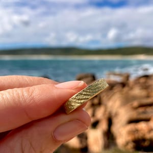 Könnte beinhalten: Ein strukturierter Goldring wird vor einem verschwommenen Hintergrund aus Strand und Meer gehalten. Der Ring hat ein einzigartiges, geriffeltes Muster auf seiner Oberfläche, das das Licht einfängt. Der Ring ist ein schlichtes Banddesign.