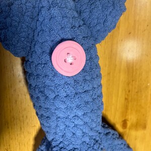 May include: A blue crochet plush toy shaped like a squid with a textured, yarn-based surface. The toy features a single, large, round, light pink plastic button attached to the center of its head, serving as a decorative eye or accent detail.