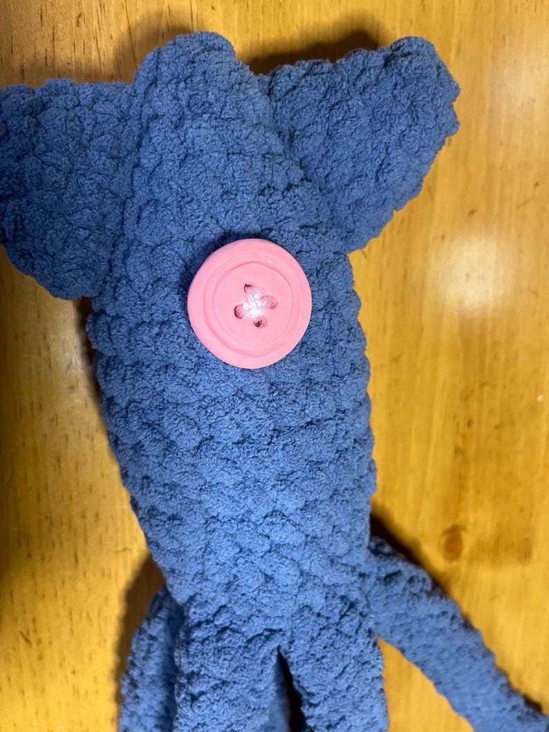 May include: A blue crochet plush toy shaped like a squid with a textured, yarn-based surface. The toy features a single, large, round, light pink plastic button attached to the center of its head, serving as a decorative eye or accent detail.
