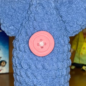 May include: A blue plush toy with a textured, looped yarn surface and a large, round, light pink button centered on the front. The toy features a wide, flat top section and a cylindrical body, showcasing a soft, tactile, and fuzzy appearance.