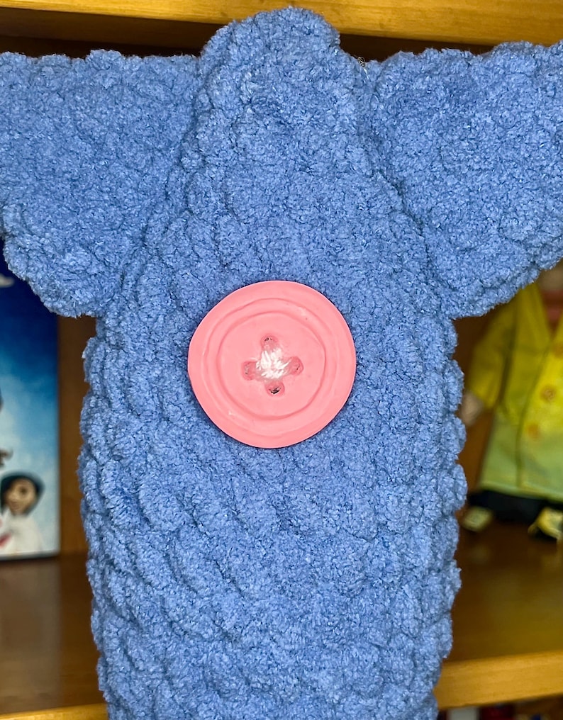 May include: A blue plush toy with a textured, looped yarn surface and a large, round, light pink button centered on the front. The toy features a wide, flat top section and a cylindrical body, showcasing a soft, tactile, and fuzzy appearance.