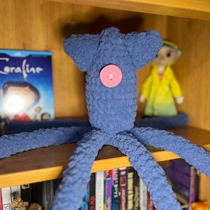 May include: A blue crocheted squid plush toy with a textured, yarn-based surface. The squid features a single, round, light pink button centered on its head, serving as a decorative detail. The plush has a wide, flat top and multiple long tentacles.
