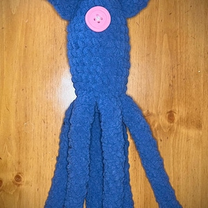 May include: A blue crocheted squid plush toy featuring a textured yarn surface, a rounded head section with two side fins, and four long dangling tentacles. A single round, light pink button is securely attached to the center of the head.