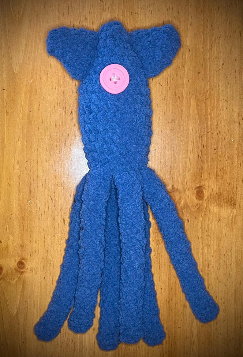 May include: A blue crocheted squid plush toy featuring a textured yarn surface, a rounded head section with two side fins, and four long dangling tentacles. A single round, light pink button is securely attached to the center of the head.